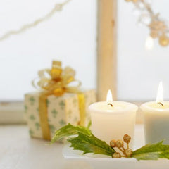 candles on a white plate with holly leaves, placed on a surface with decorative lights in the background.