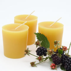 Three yellow candles with wicks on a white background, accompanied by blackberries and leaves.