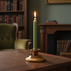 Green candle in an amber glass holder on a wooden surface with a bookshelf and chair in the background.