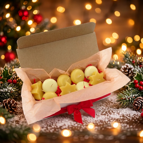 An open kraft gift box lined with blush pink tissue paper, filled with small round and star-shaped yellow candles, tied with a red ribbon. The box is styled on a snowy surface with pine branches, pinecones, and warm holiday bokeh lights in the background.