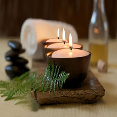 Three lit candles in black bowls on a wooden surface with fern leaves and stones in the background.