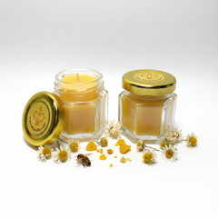 Two hexagonal glass jars filled with golden yellow honey, one open with its gold lid leaning beside it and one closed with a gold lid. Small daisy flowers, loose honey droplets, and a small bee decoration are arranged around the jars on a clean white background.
