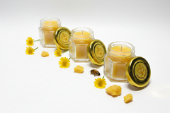Three hexagonal glass jars filled with yellow wax candles, each with a gold lid, arranged on a white background with scattered yellow flowers, wax chunks, and a small bee.