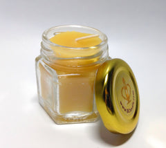 A small hexagonal glass jar candle filled with pale yellow wax, shown open with its gold-colored lid leaning against it on a white background.