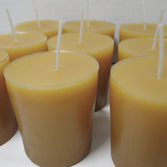 A group of unlit beeswax candles, showing a warm tan wax color with white wicks, arranged closely together on a light surface